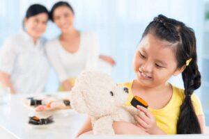 A child pretending to feed cake to a teddy bear while adults watch in the background